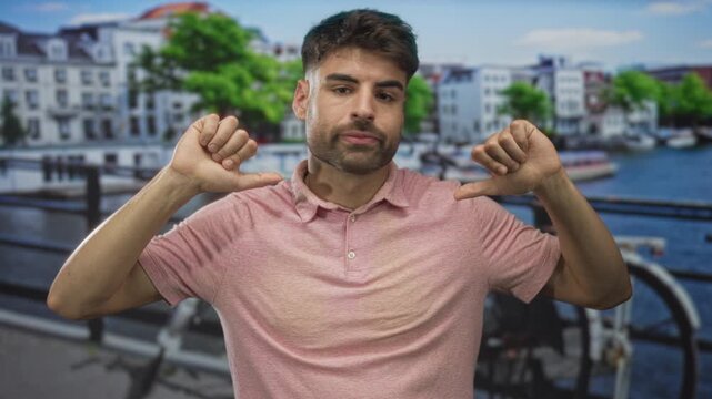Young hispanic man in pink shirt closing eyes and giving double thumbs down on a street; disapproval.