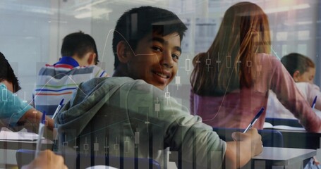 Smiling teenage boy in gray hoodie holding pen above paper at classroom desks, with notebooks