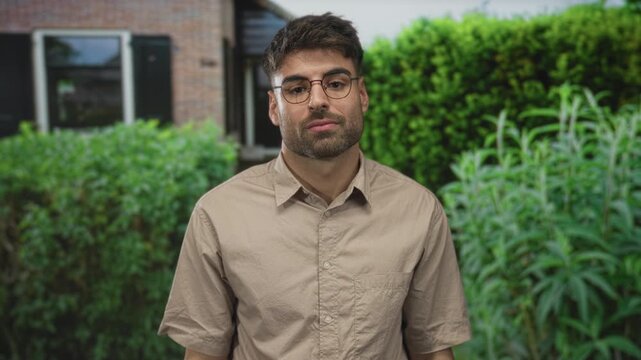 Man in beige shirt shrugs shoulders at building entrance framed by lush green hedge; inner uncertainty.