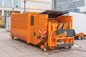 A large orange industrial waste compactor stands on a city square. The machine has visible hydraulic arms and safety markings. © Jakob