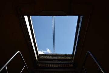 Looking up at a clear blue sky through a dark, rectangular hatch, with two parallel wires bisecting the frame
