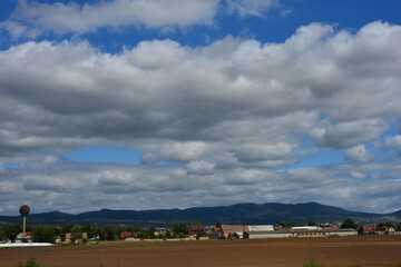 A rural landscape with a plowed field in the foreground, a small town, and a water tower at the base of distant mountains