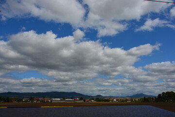 A dramatic, cloudy blue sky stretches over a small town nestled at the base of a long, dark range of forested hills