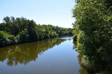 A calm, wide river winds its way through a dense green forest, with the trees reflected on the water under a clear blue sky