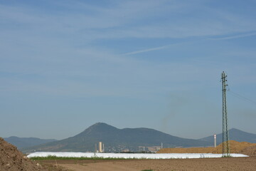 An industrial and agricultural landscape with a power line, factory chimneys, and silage bags against a backdrop of distant mountains