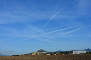 A rural landscape with agricultural buildings and a plowed field under a vast blue sky marked by numerous crossing airplane contrails