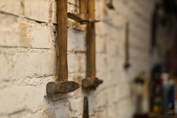 Several old, rustic hammers with wooden handles hang vertically on a textured white brick wall in a workshop.