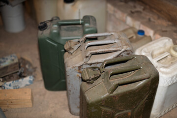 Several old and dusty jerry cans, made of metal and plastic, are arranged on a concrete floor in a dimly lit storage area.