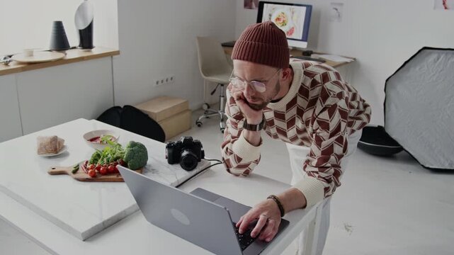 High angle shot of Caucasian man working on laptop, sorting photos. He surrounded by props and photography equipment