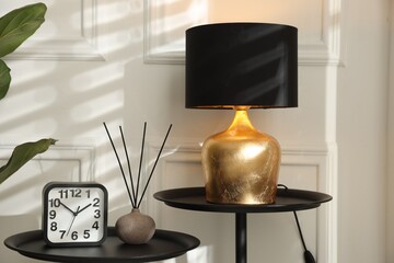 Stylish lamp, reed air freshener and alarm clock on black tables near white wall indoors