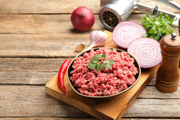 Raw minced meat with parsley in bowl, grinder and products on wooden table. Space for text