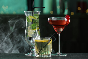 Glasses of tasty cocktails on dark table against blurred background with smoke, bokeh effect