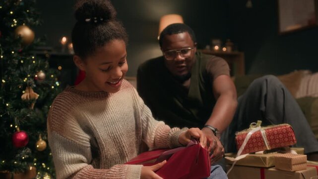 Joyful Black girl child opening wrapped gift box receiving Christmas present from loving father while sitting on floor in decorated living room in evening