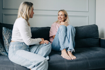 Two smiling beautiful blonde women are chatting while sitting on the sofa in the living room.