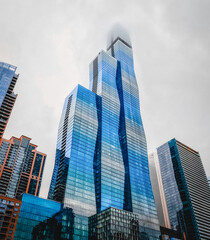 Obraz premium Majestic glass skyscraper buildings reach to a cloudy sky on the Chicago River in Chicago, Illinois. The curvy blue skyscraper is the third tallest building in Chicago. Located in the Loop.