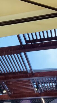 Diagonal view from beneath a cream awning as rain falls in bright streaks. Drops splash off a timber pergola and guttering, with pale sky beyond. Moody, cozy patio moment
