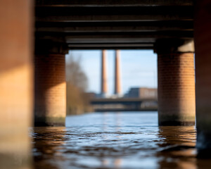River view under bridge: Waters flow between brick pillars, revealing distant industrial chimneys. Urban scene merges nature and architecture.