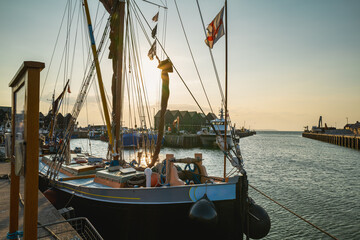 Golden evening as the setting sun is reflected the water of Whitstable harbour behind the impressive mast and rigging of a thames barge moored in the foreground and in front of Fishermans huts.