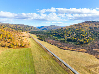 Wide aerial view of a straight paved road dividing a valley of agricultural fields and distant hills covered in yellow and green autumn forest