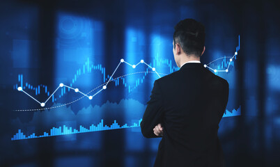 A business professional stands with his back to the camera, observing digital graphs and charts in...