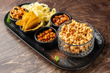 Assorted party snacks in black tray on wooden table. Includes popcorn, chips, pretzels, cashews, chickpeas, puffed rings. Top view, neat presentation, garnished with parsley.