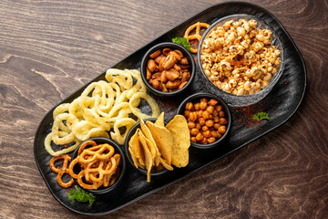 Assorted party snacks in black tray on wooden table. Includes popcorn, chips, pretzels, cashews, chickpeas, puffed rings. Top view, neat presentation, garnished with parsley.