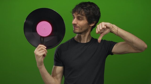 Man holds vinyl record with pink label and shows a thumbs down gesture in a vibrant green studio; disapproval.