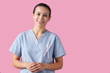 Portrait of young female doctor smiling and wearing medical scrubs with pink ribbon pinned to chest, standing against pink background representing breast cancer awareness