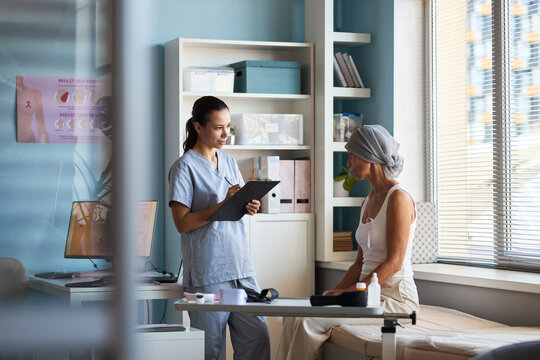 Middle aged Caucasian woman wearing headscarf sitting on examination table listening to young adult female doctor holding clipboard discussing cancer diagnosis in medical office