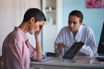Obraz premium Middle aged Caucasian woman sitting with hand on forehead, showing distress, while female doctor explaining diagnosis using digital tablet, in medical office discussing cancer treatment
