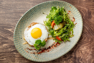 Gourmet hamburger patty topped with sunny-side-up egg, creamy sauce, and parsley, served with fresh salad on elegant plate. High-end food presentation, rustic wooden background.
