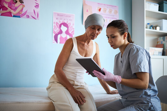 Middle aged Caucasian woman with headscarf sitting on examination table consulting with young adult Caucasian female healthcare worker holding digital tablet in medical office