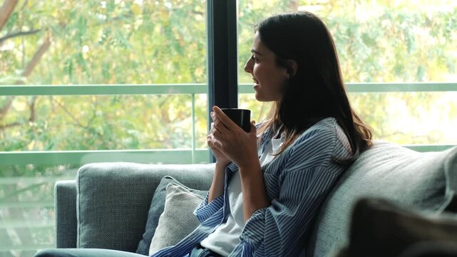 Young woman sitting on sofa at home, smiling and relaxed while drinking a cup of tea, coffee or herbal infusion, symbol of calm, comfort, wellbeing, leisure and cozy lifestyle