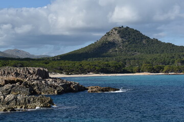 Cala Agulla Mallorca with turquoise waters rugged rocks and lush green forest at the base of a scenic mountain under a partly cloudy sky
