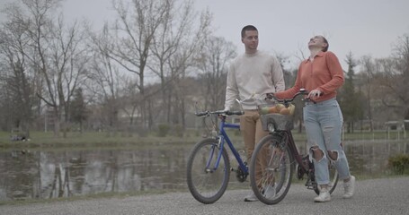 A happy couple enjoys a leisurely walk with their bicycles beside a tranquil lake in a beautiful park. They share smiles and conversation, embracing nature and healthy outdoor activity together. - Powered by Adobe