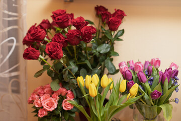 A vibrant display of red roses, yellow tulips, and pink tulips arranged in bouquets against a light wall and sheer curtain.