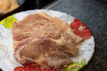 Raw, breaded meat cutlets are arranged on a white plate with colorful flower designs, ready for frying.