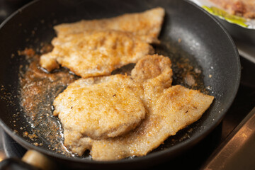 Several pieces of breaded meat are frying in a black pan, showing a golden-brown crust and...