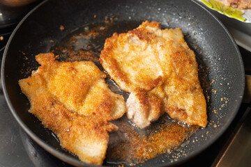 Two golden-brown breaded cutlets are sizzling in a black frying pan, surrounded by oil and cooking crumbs.