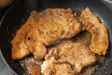 Several breaded meat cutlets are pan-frying in oil, developing a golden-brown crust with visible seasoning in a dark skillet.