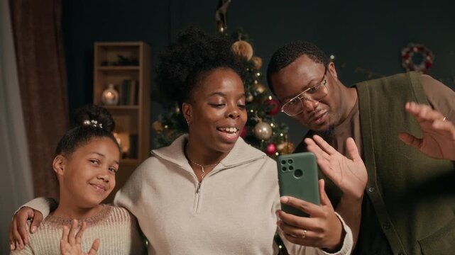 Handheld shot of cheerful Black woman with girl child and husband chatting with relatives in video call waving hands and showing Christmas tree in decorated living room in evening