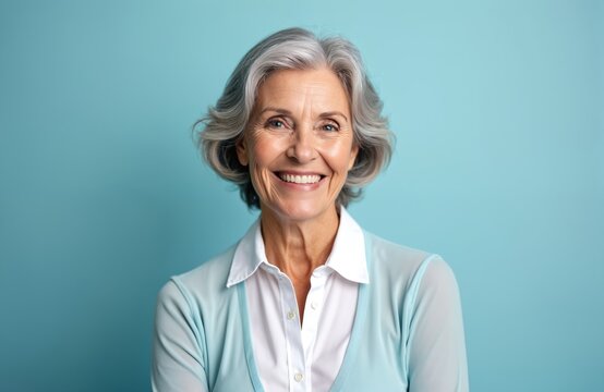 Smiling senior woman looks into camera with bright, friendly expression. Grey hair styled neatly, wears light blue cardigan over white blouse. Radiates warmth, contentment against solid blue