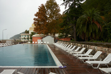 A panoramic view of the hotel's waterfront on a rainy day. The pool is covered in raindrops and the sun loungers are wet. A building fragment is visible in the background.