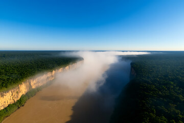 Fototapeta premium Aerial view of a wide river cutting through a dense forest, partially obscured by mist, under a clear blue sky, creating a serene landscape.