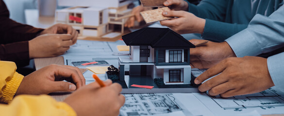 A diverse architectural team engages in a creative session, examining a detailed scale model of a house alongside blueprints and planning materials on a modern workspace table. SACTR