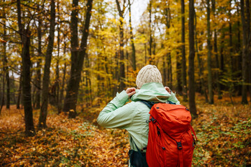 Female tourist in sports clothing with backpack hiking in autumn forest. Woman hiker wearing waterproof jacket with hood.