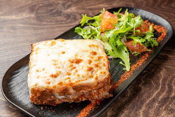 Baked lasagna with melted cheese served on a black plate with fresh arugula salad, cherry tomatoes, avocado, and paprika garnish on rustic wooden background.