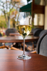 A glass of white wine sits elegantly on a wooden table, surrounded by trees and outdoor seating