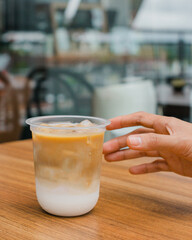 Iced latte in a plastic cup on a wooden table with a hand reaching for it.