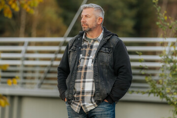 Man in a plaid shirt and denim jacket stands outdoors near a metal railing, surrounded by soft autumn tones and warm natural light.